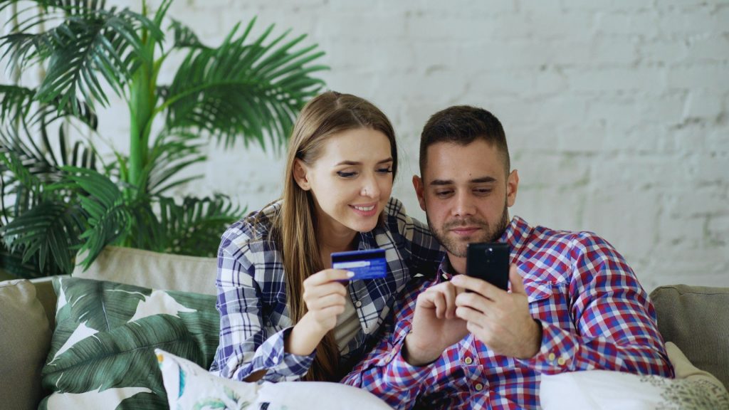 Un couple regarde un smartphone, carte bancaire en main, sur un canapé